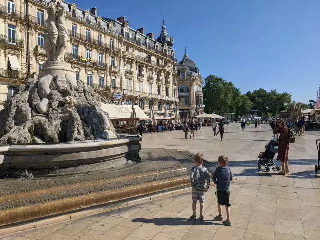 Two young boys look at a large stone fountain in a sunny French plaza lined with ornate buildings and cafes