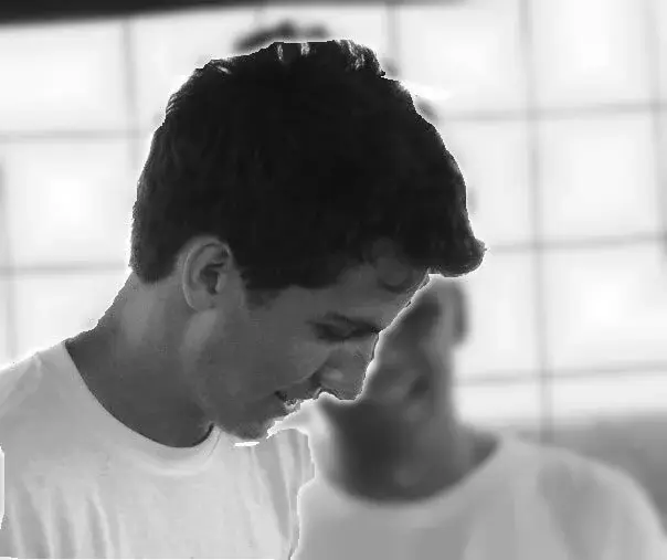 Black and white photo of a young man in a white T-shirt looking down, with another person blurred behind him