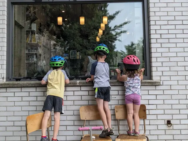 Three kids in bike helmets stand on wooden chairs, peering into a cafe window set in a white brick wall